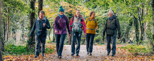 A mixed-age group of people walk together along a woodland path in autumn, wearing outdoor jackets and backpacks, with one adult carrying a baby in a front carrier as they chat and smile among fallen leaves.